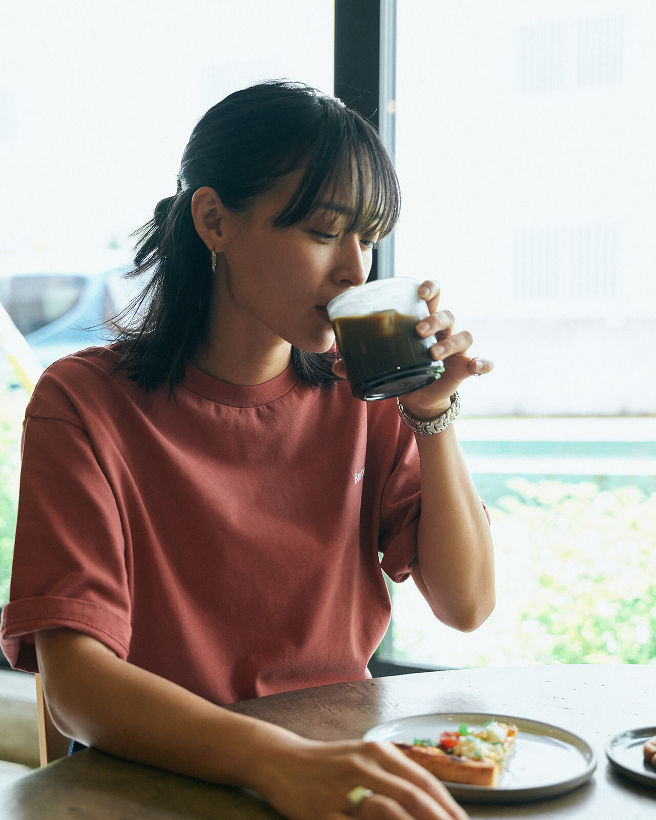 ピンクのトップス・ブルーデニムのスカートを着用した女性が席に座ってコーヒーを飲んでいる様子