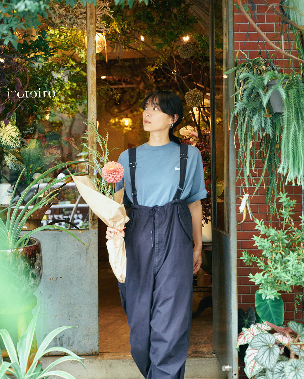 ライトブルーのトップス・ネイビーのボトムスを着用した女性がお花を持ちながらお花屋さんを出る様子