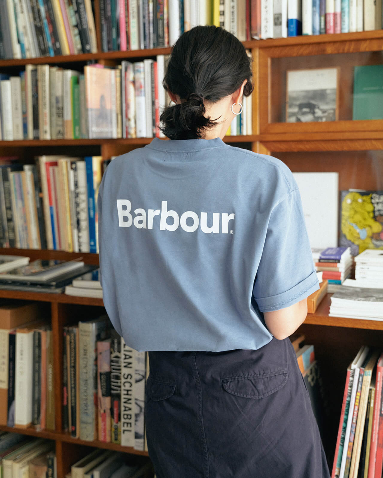 ライトブルーのトップス・ネイビーのボトムスを着用した女性が本屋さんで本を立ち読みしている後からの様子